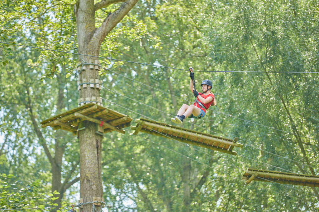 Vrouw in rood shirt met zwarte helm ziplined langs andere parcoursen.