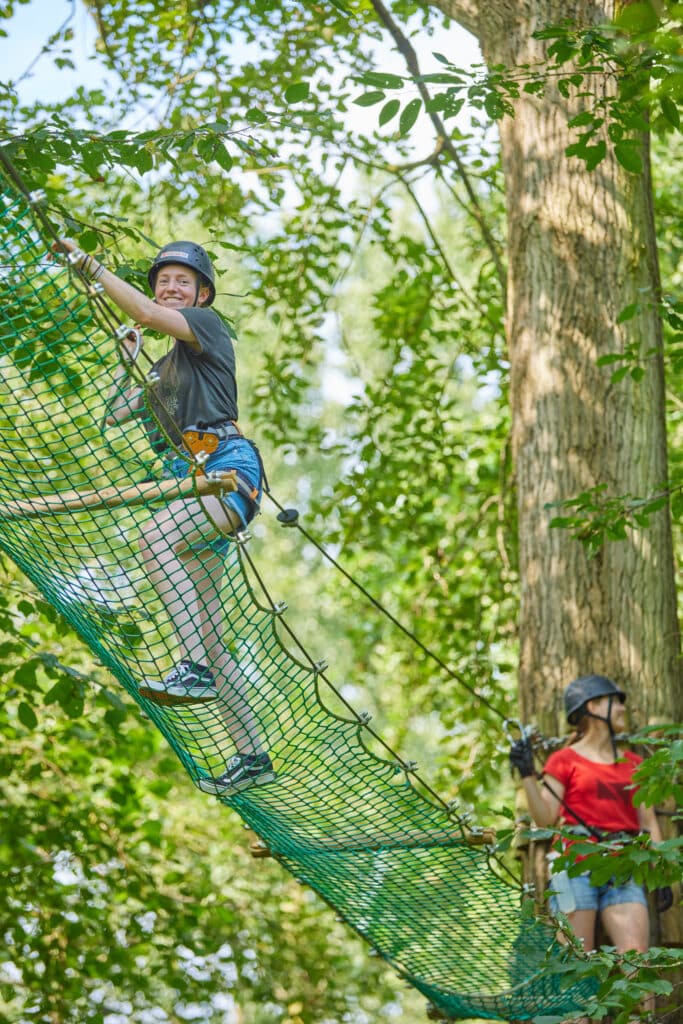 Jongvolwassene met zwarte helm en zwart shirt over een net dat omhoog loopt. Op de achtergrond staat een vrouw in een rood shirt te wachten tot zij mag klimmen.