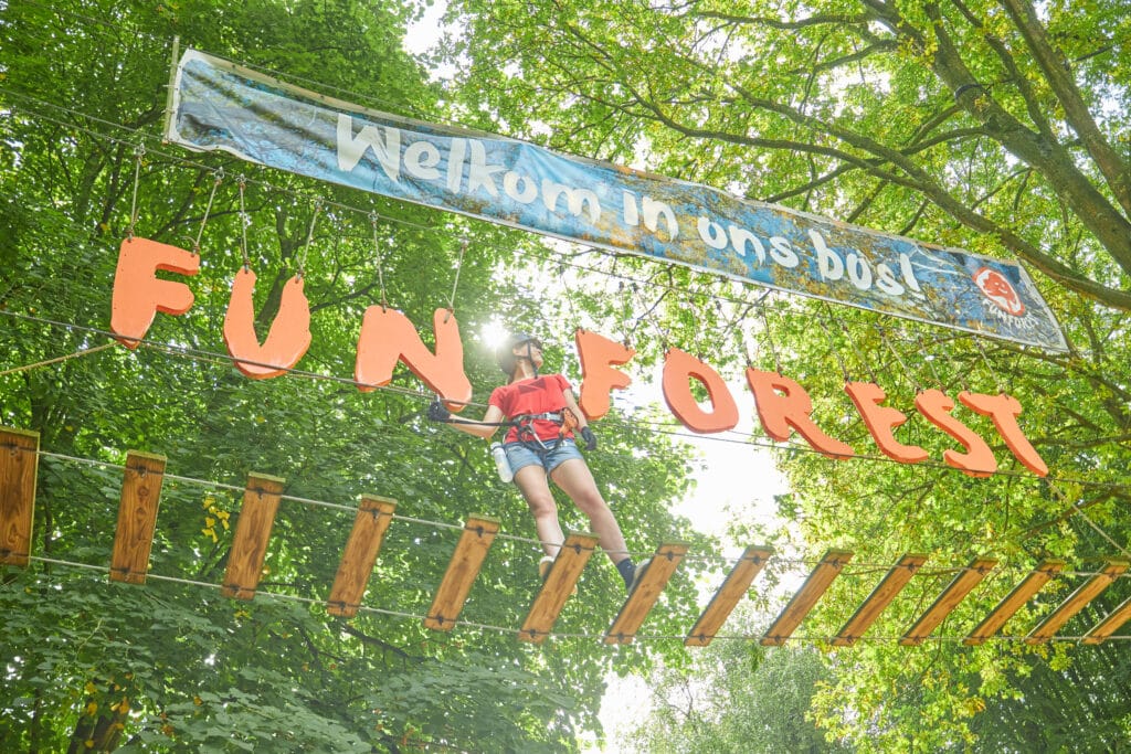 Vrouw staat met een glimlach op het gezicht en kijkend naar rechts voor de letters 'Fun Forest'. Boven de letters hangt een grote banner met 'Welkom in ons bos!'.