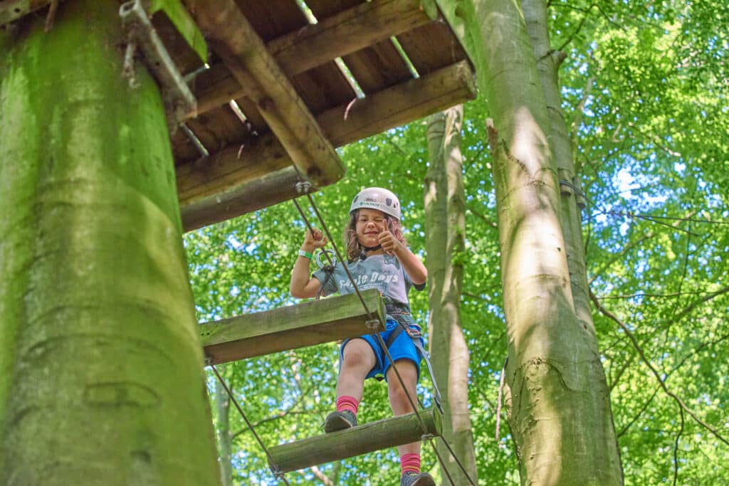 Jongetje steekt zijn duim op en knipoogt naar de camera terwijl hij tussen de bomen over een brug met houten palen klimt.