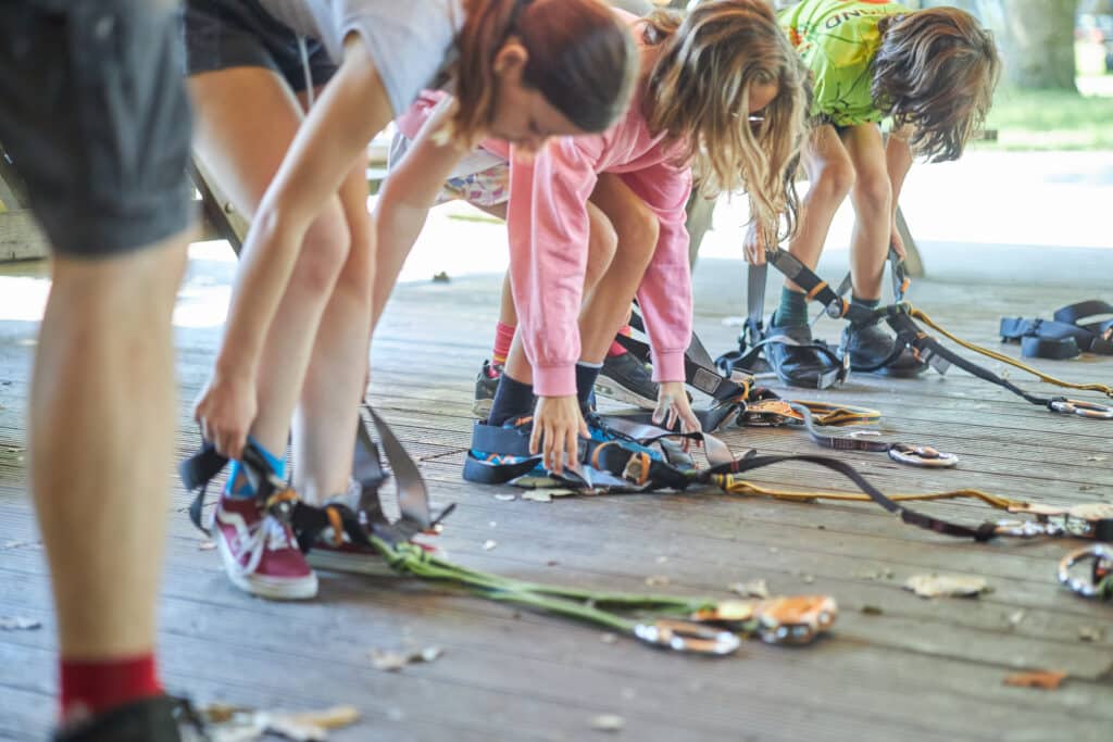 Drie kinderen trekken hun gordels aan. De gordels hebben twee beenlussen en twee lifelines waar een zaza en een katrol aan vastzit.