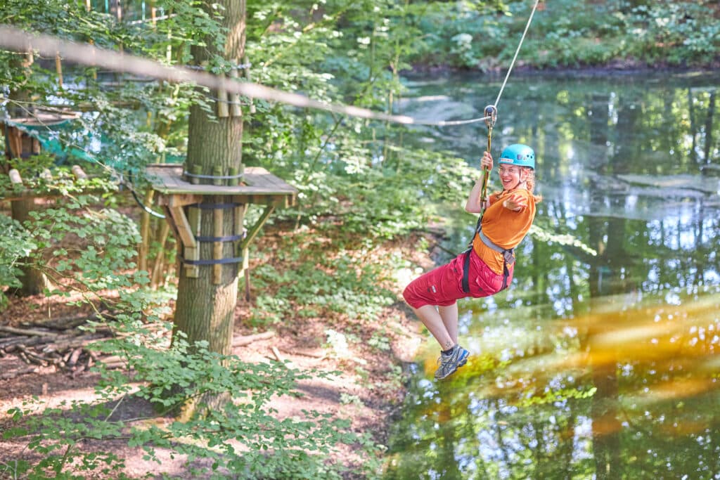 Vrouw tokkelt naar de overkant van het water. Ze heeft een blauwe helm op haar hoofd en strekt haar arm uit naar de camera.