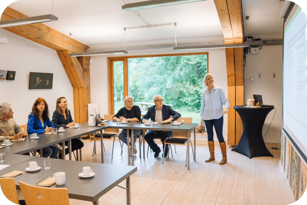 6 mensen zitten in een vergaderruimte. De tafels staan in een u-vorm, boven op de tafels staan lege witte kopjes. 1 persoon staat naast de tafels en refereert aan het scherm wat tegenover de tafels hangt.